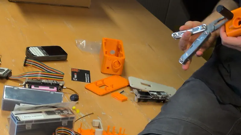 Hands working on an orange prototype case above a bench with the open enclosure, circuit boards, battery and wiring laid out.