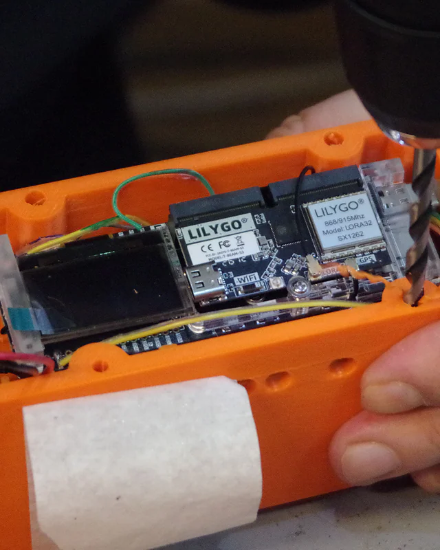 Hands holding the orange MSC-2 case while a drill works on it, the board already fitted inside with its display and LoRa module visible.