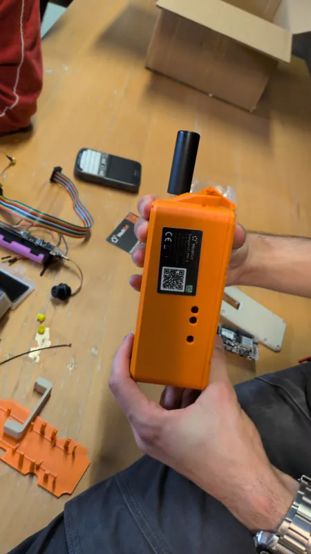 Orange printed MSC-2 prototype enclosure held in two hands, with its antenna and rear product label visible; loose parts remain on the workbench.