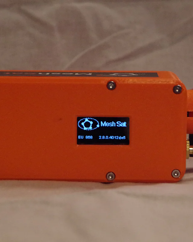 The finished prototype unit 0001 lying on a cloth, its display showing the MeshSat start screen, with a red USB cable and the antennas at the ends.