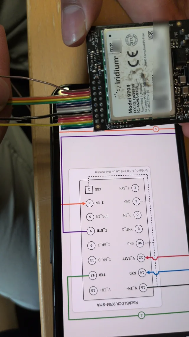 The 9704 modem held above a phone showing the MSC-2 wiring sheet, the six-wire ribbon harness soldered to its header being checked pin by pin.