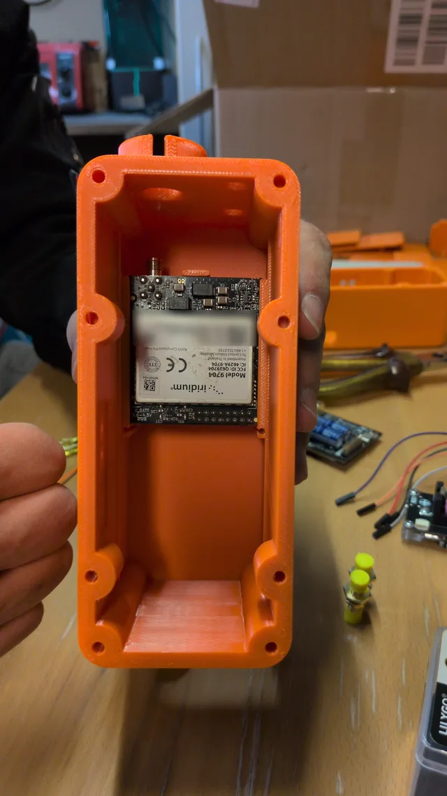 The RockBLOCK 9704 satellite modem lying in the lower bay of the open orange MSC-2 case, its antenna socket toward the end wall, held over a workbench.