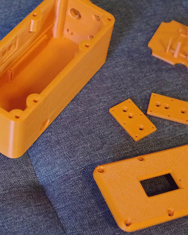 The orange printed parts of an MSC-2 enclosure laid out on a blue sofa before assembly: the case, the lid with its display window, the inner bracket and two small mounting plates.